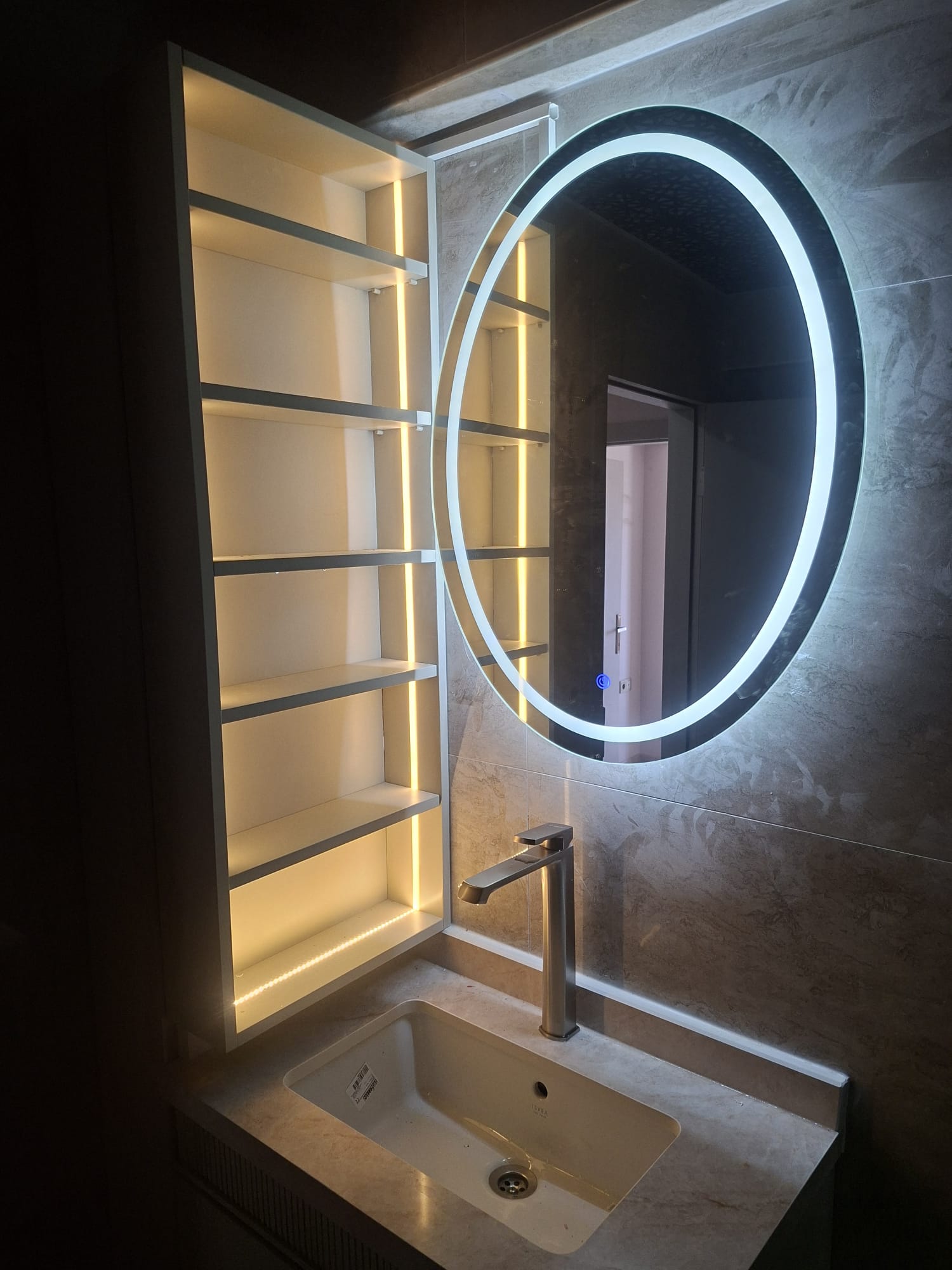 Yuvarlak ışıklı ayna, açık raf ve taş dokulu banyo yüzeyleri